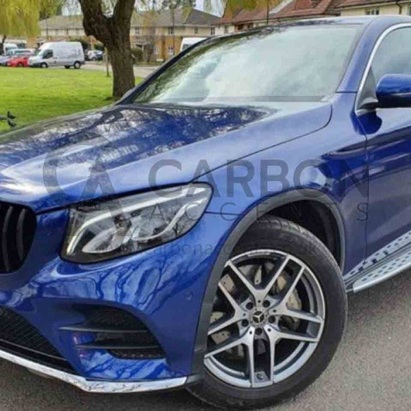 A blue Mercedes car parked on a road with a side view of large alloy wheels. Background features a grassy area with trees and buildings. Text: "CARBON ACCENTS" on the license plate.