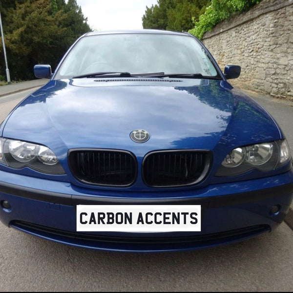 A blue sedan is parked on a street next to a stone wall. The license plate reads "CARBON ACCENTS." The vehicle's emblem is modified, surrounded by greenery and pavement.