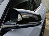 A sleek, black car side mirror reflects clouds, attached to a silver vehicle in an outdoor setting with a corrugated metal fence in the background.