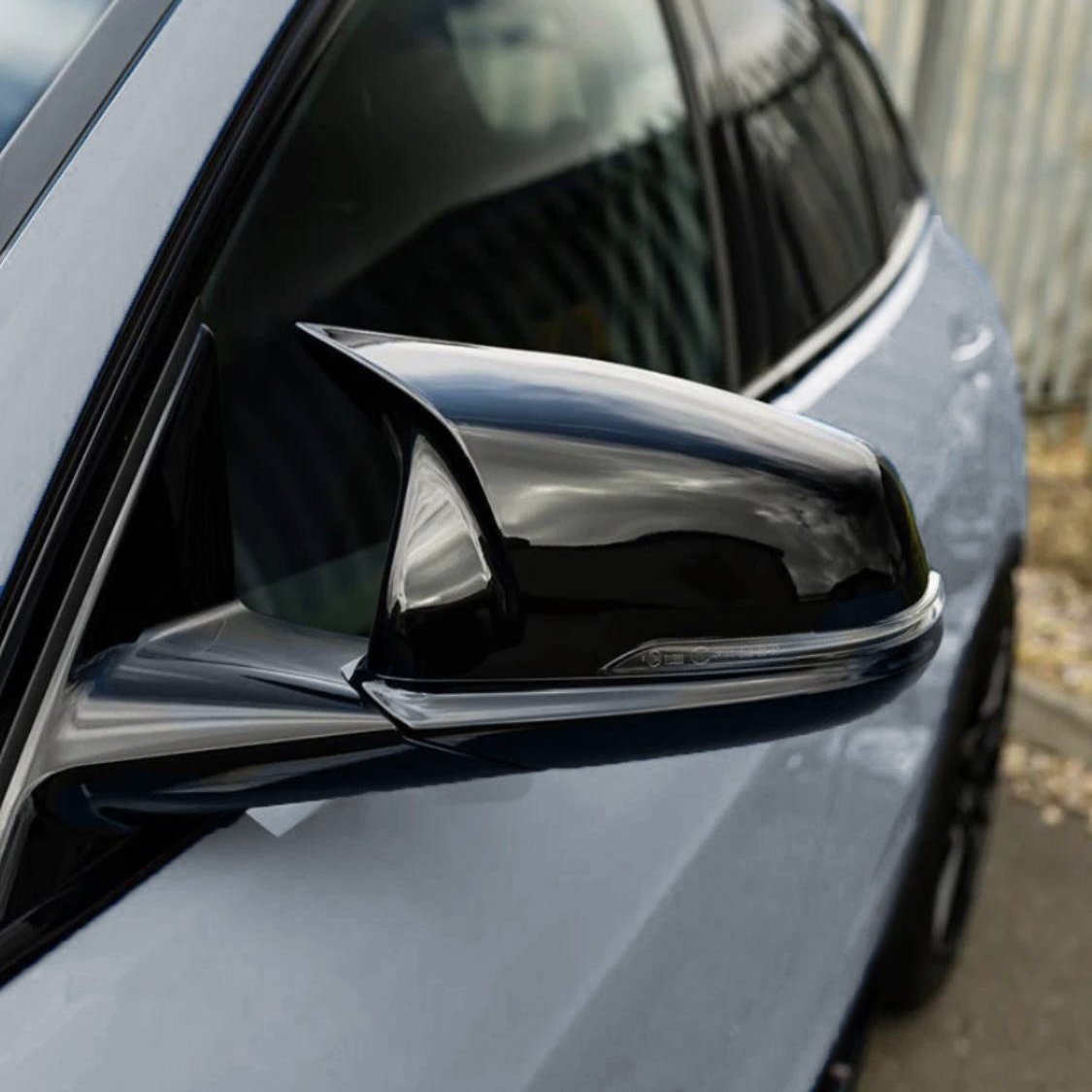 A sleek, black car side mirror reflects clouds, attached to a silver vehicle in an outdoor setting with a corrugated metal fence in the background.