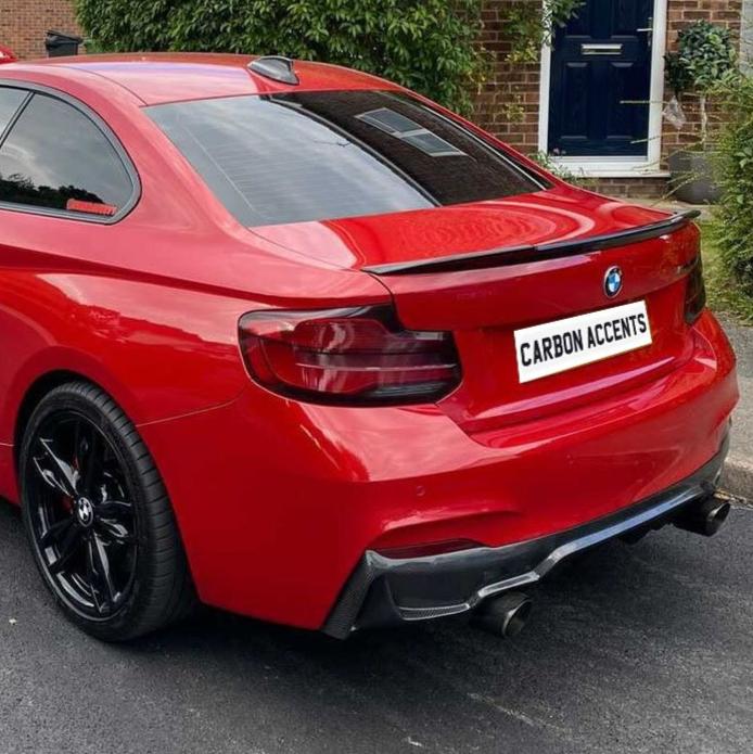 A red BMW car parked on a street, displaying the license plate "CARBON ACCENTS," against a backdrop of a brick house with a blue door and surrounding greenery.