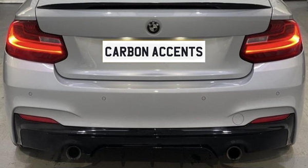 A white BMW car's rear, illuminated taillights, in a garage setting. License plate reads "CARBON ACCENTS." The car has a black lower bumper and dual exhaust pipes.