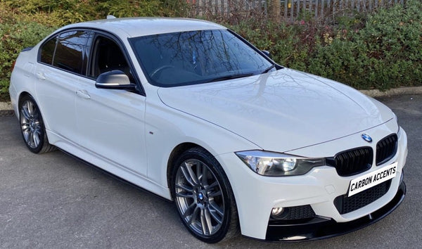 White sedan parked on asphalt, featuring sleek carbon accents and a front license plate reading "CARBON ACCENTS"; surrounded by greenery and a metal fence.