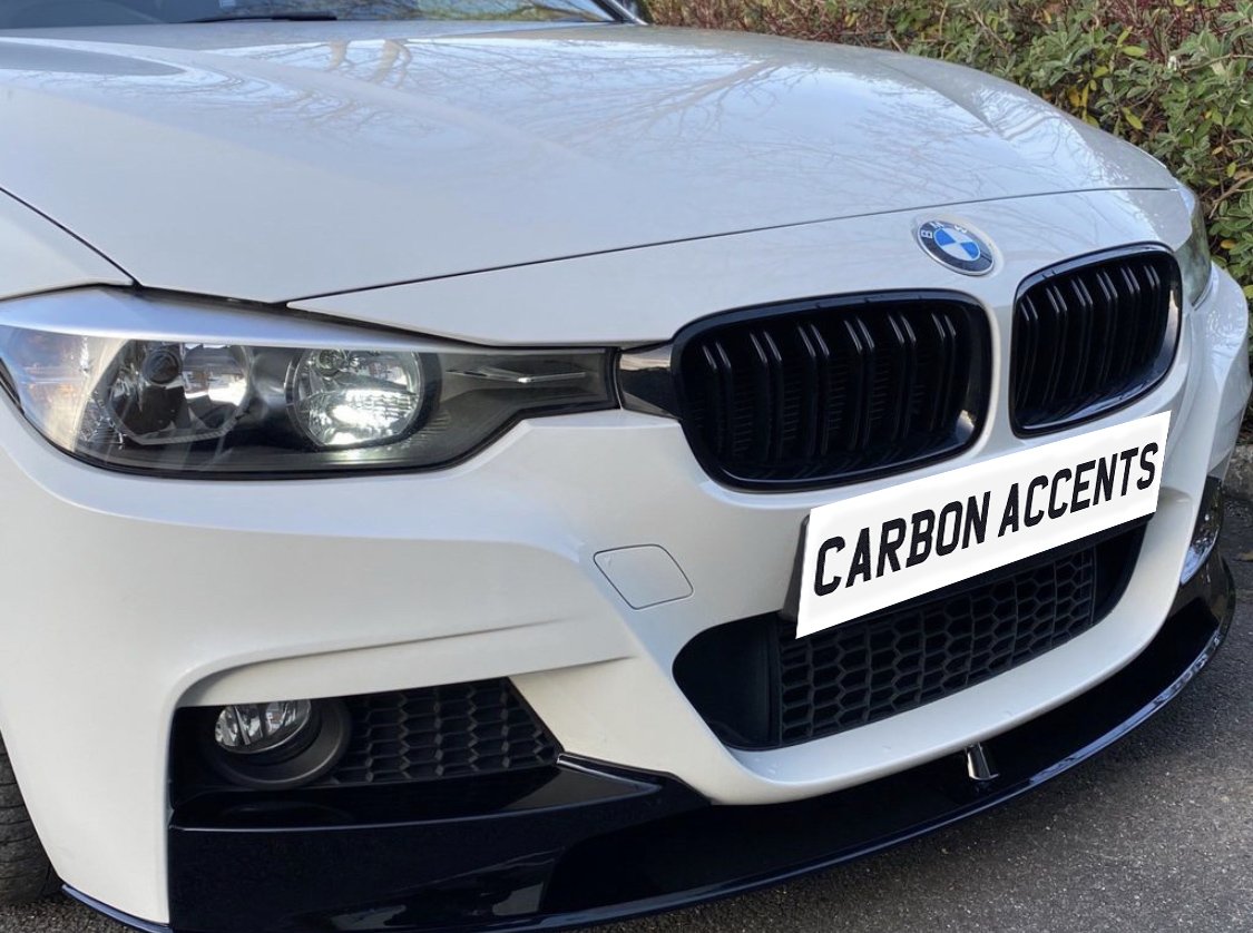 A white BMW car front with illuminated headlights, featuring a black grille. The license plate reads "CARBON ACCENTS." It's parked outdoors near bushes.