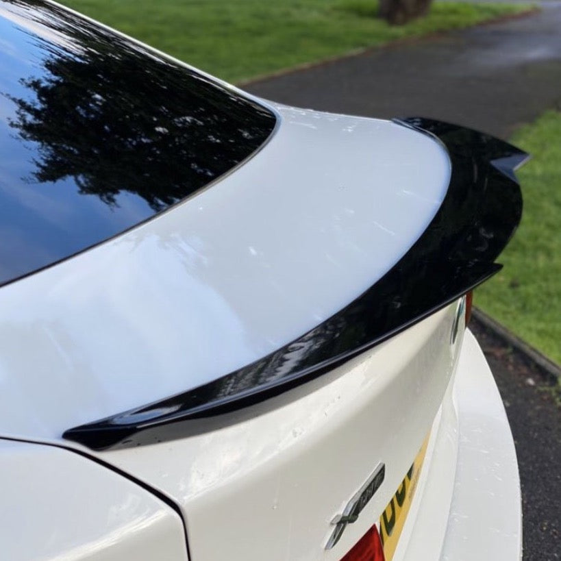 A white car with a black spoiler is parked on a paved path beside a grassy area. Reflection of trees is visible on the car's surface. The license plate shows "007".