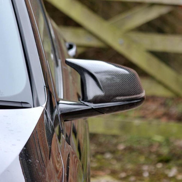 Car side mirror with carbon fiber casing reflecting the car's sleek body, set against a blurred wooden fence background.