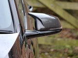 A close-up of a car's carbon fiber side mirror reflects a blurred background of wooden fencing and nature, indicating an outdoor setting.