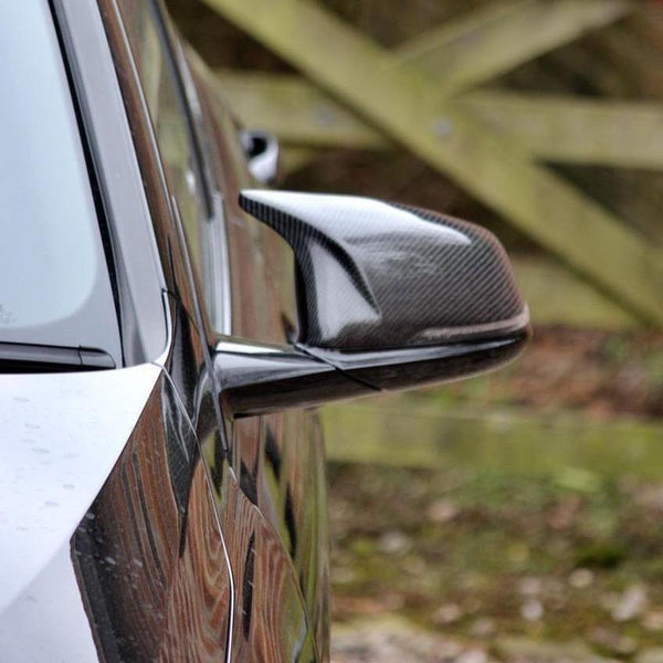 A close-up of a car's carbon fiber side mirror reflects a blurred background of wooden fencing and nature, indicating an outdoor setting.