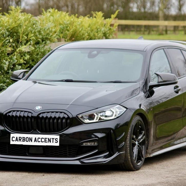 A black car is parked on a driveway, surrounded by greenery. The license plate reads "CARBON ACCENTS."