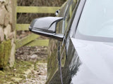 A black car's side mirror reflects light while parked beside a moss-covered stone wall and wooden fence on a gravel path.