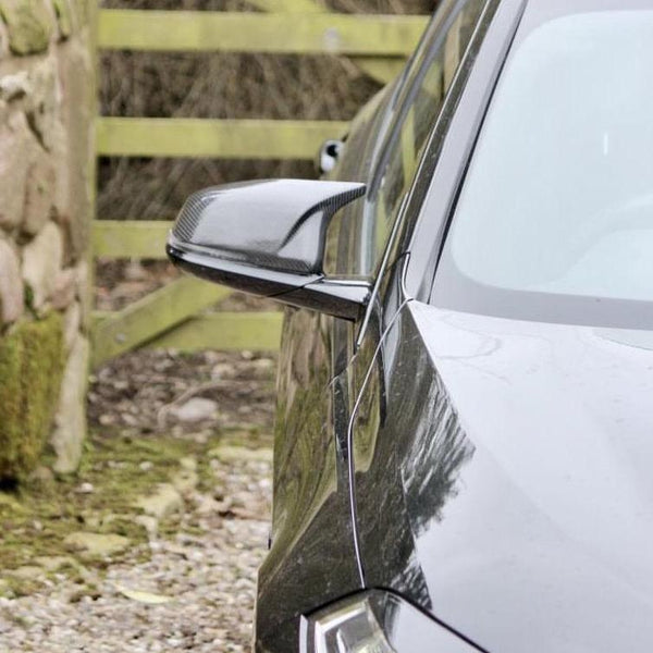 A black car's side mirror reflects light while parked beside a moss-covered stone wall and wooden fence on a gravel path.