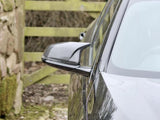 A sleek car's side mirror is shown outside near a moss-covered stone wall and a wooden gate in a rural setting.