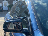 A blue car's side mirror reflects nearby buildings on a sunny street, with brick houses and white doors visible in the background.