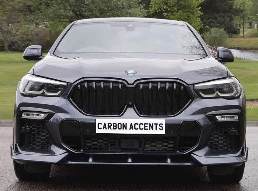 A sleek, black car with a prominent grille and sharp headlights is parked on a driveway. The surroundings feature grassy lawns and trees. Text on the grille reads "CARBON ACCENTS."