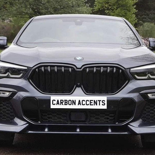 A sleek, black car with a prominent grille and sharp headlights is parked on a driveway. The surroundings feature grassy lawns and trees. Text on the grille reads "CARBON ACCENTS."