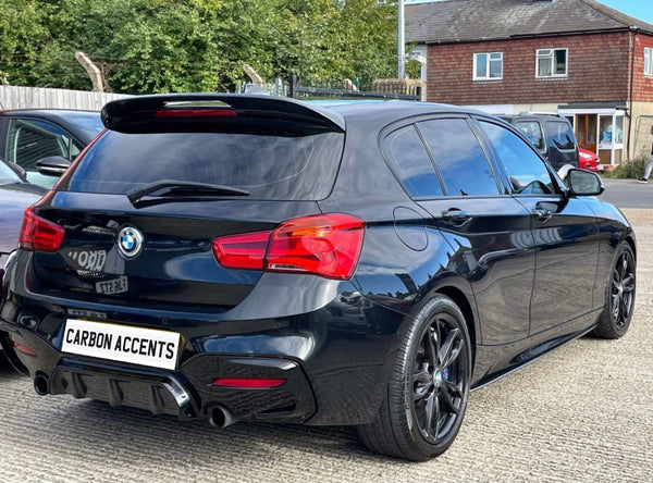 A black BMW hatchback is parked on a concrete surface, featuring tinted windows and red taillights. The car's license plate reads "CARBON ACCENTS," with trees and buildings in the background.