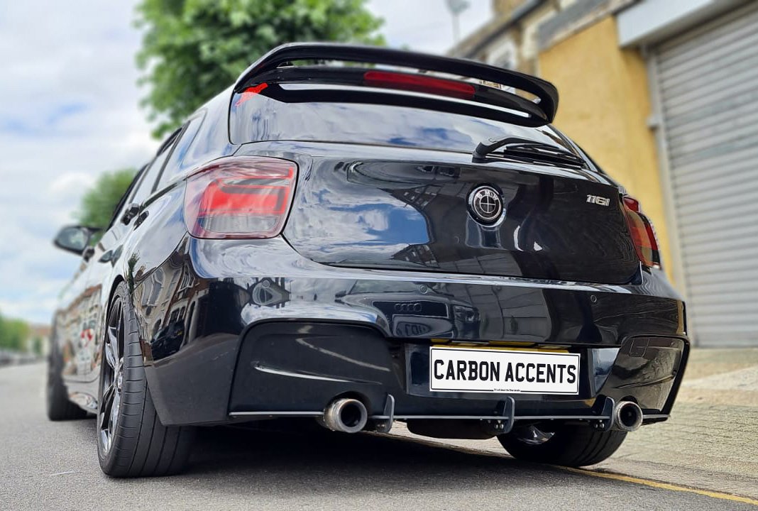 A sleek black car with a rear spoiler is parked on a street. It has dual exhaust pipes and a license plate reading "CARBON ACCENTS." Nearby, buildings and trees border the road.