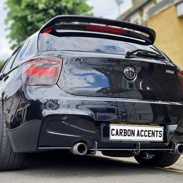 A sleek black car with a rear spoiler is parked on a street. It has dual exhaust pipes and a license plate reading "CARBON ACCENTS." Nearby, buildings and trees border the road.