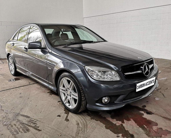 A dark gray sedan, parked on a concrete surface, has reflective windows and alloy wheels. The visible license plate reads "CARBON ACCENTS." The background features white brick walls.