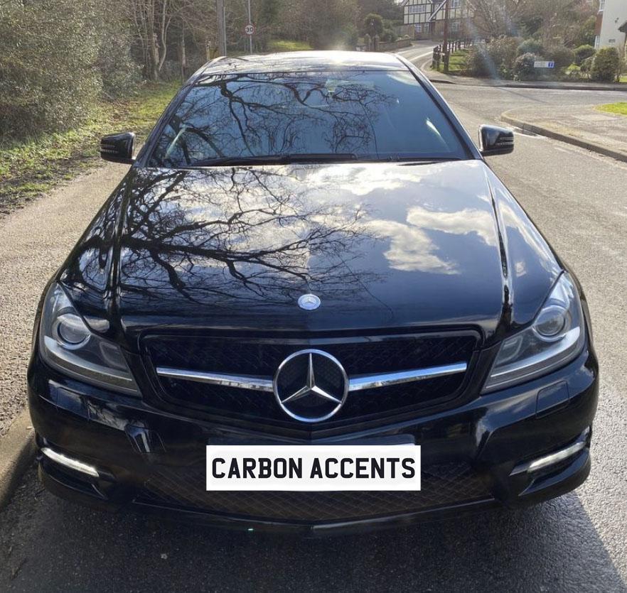 A black Mercedes car is parked on a street, reflecting surrounding trees on its shiny hood. The license plate reads "CARBON ACCENTS." The street has greenery on the side.