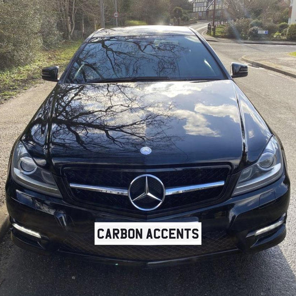A black Mercedes car is parked on a street, reflecting surrounding trees on its shiny hood. The license plate reads "CARBON ACCENTS." The street has greenery on the side.