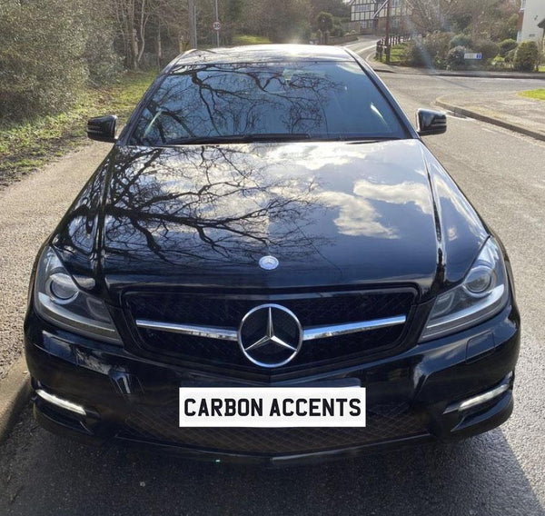 A black Mercedes car is parked on a street, reflecting surrounding trees on its shiny hood. The license plate reads "CARBON ACCENTS." The street has greenery on the side.