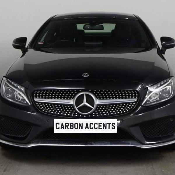 A black Mercedes-Benz car is parked indoors, facing front. Its grille features the Mercedes emblem. Below, a plate displays the text "CARBON ACCENTS." The background is plain white.