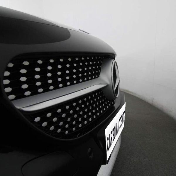 A car's front grille features a prominent logo with a pattern of hexagonal elements. It is parked in a well-lit garage. The visible text reads "CARBON ACCENTS."