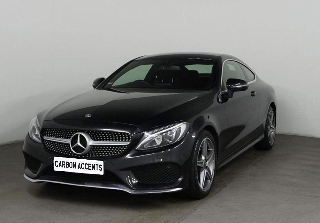 A sleek black Mercedes-Benz coupe is parked in a plain, well-lit showroom. The front license plate reads "CARBON ACCENTS," and the car features shiny alloy wheels.