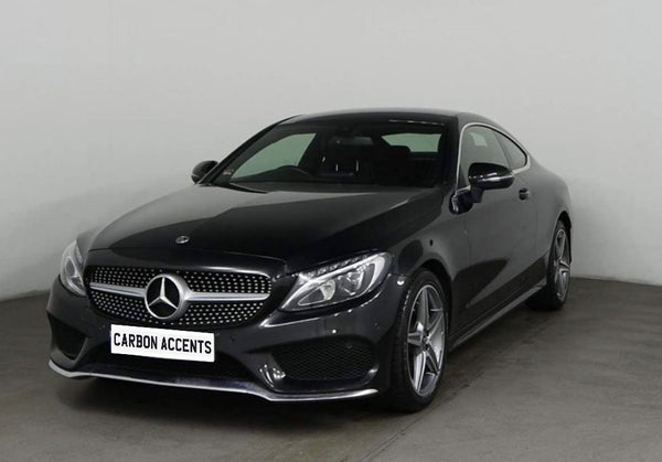 A sleek black Mercedes-Benz coupe is parked in a plain, well-lit showroom. The front license plate reads "CARBON ACCENTS," and the car features shiny alloy wheels.