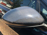 A car side mirror with a carbon fiber pattern is attached to a black vehicle. It reflects nearby houses and a tree on a sunny day, showing a suburban street scene.