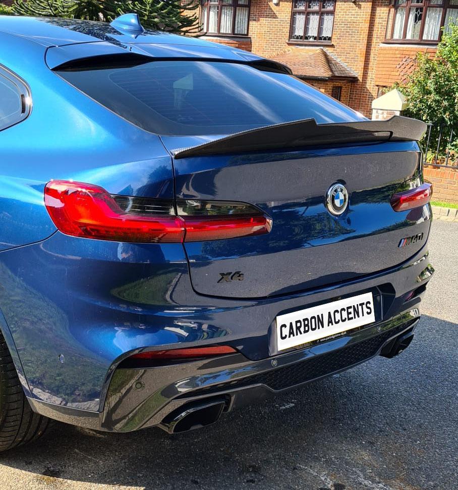 A blue BMW X6 car, parked on a street, features a spoiler and twin exhausts. The license plate reads “CARBON ACCENTS.” Brick houses and greenery surround the vehicle.