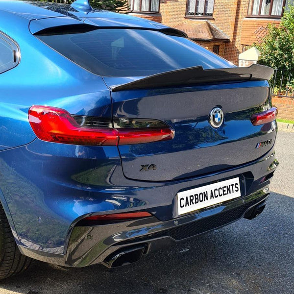 A blue BMW X6 car, parked on a street, features a spoiler and twin exhausts. The license plate reads “CARBON ACCENTS.” Brick houses and greenery surround the vehicle.