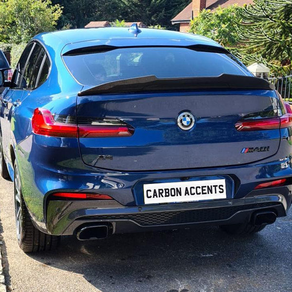 BMW car parked on a residential street with greenery visible. The license plate reads "CARBON ACCENTS." Nearby, there's another vehicle and houses in the background.