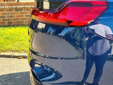 A blue car's rear with a red taillight is parked on a street. A person's reflection is visible on the car. A brick house and grass are in the background.