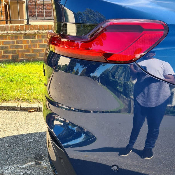 A blue car's rear with a red taillight is parked on a street. A person's reflection is visible on the car. A brick house and grass are in the background.