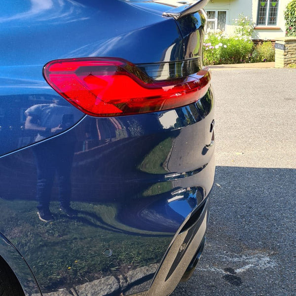 A blue car with a visible taillight and spoiler is parked on a sunny street, reflecting a residential house and greenery in its shiny surface.