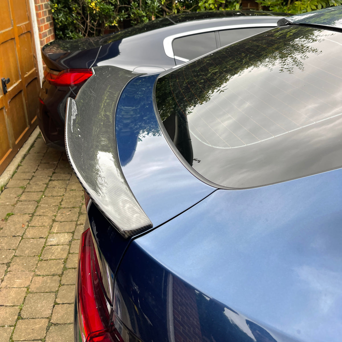 A blue car with a carbon fiber spoiler is parked on a brick driveway next to a black car, surrounded by a wooden gate and green foliage.